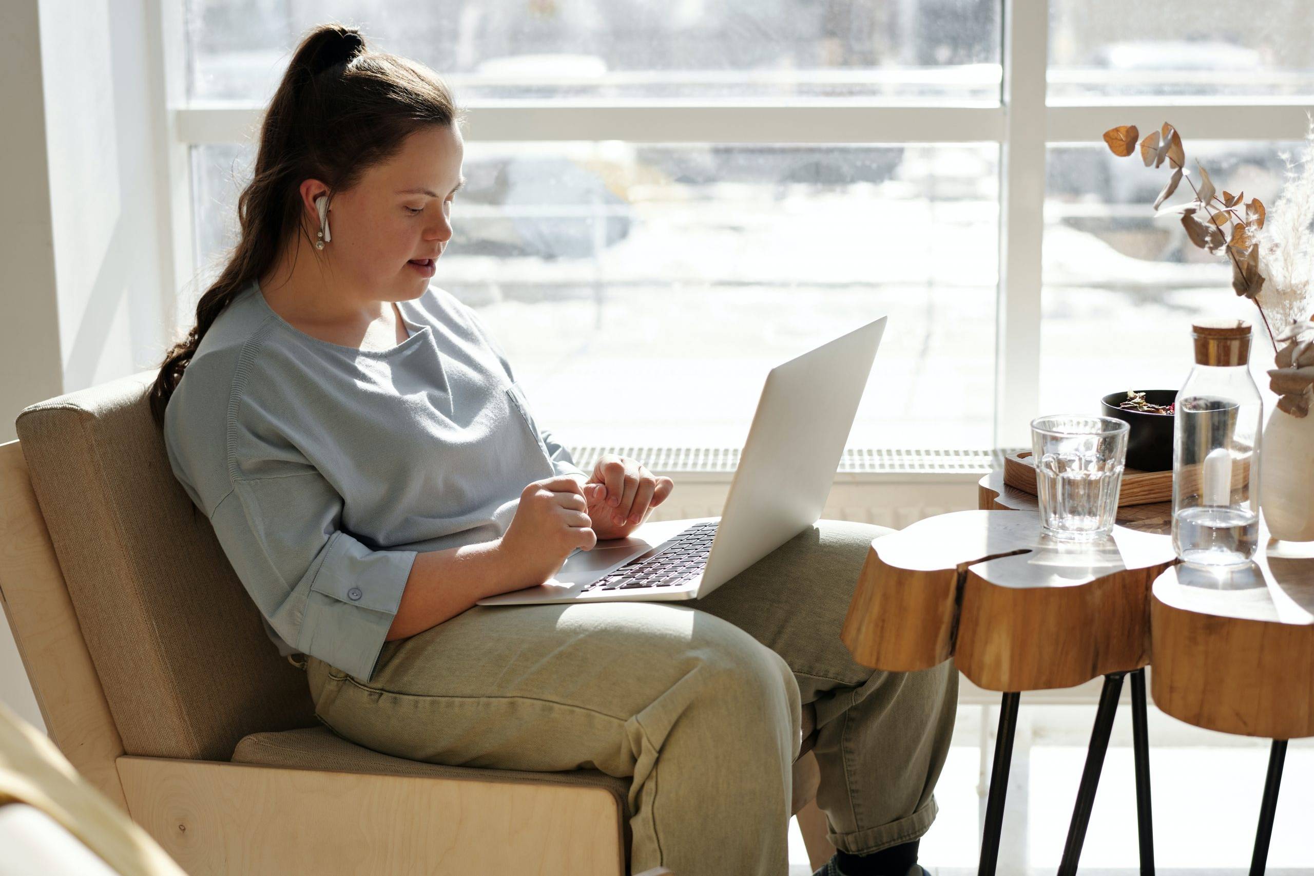 A woman with down's syndrome sitting on a beige chair without arm rests. She has brown hair which is tied up in a high ponytail, she is wearing airpods and a light blue long sleeve three quarter length top, beige loose trousers and has a laptop on her lap. There is a wooden table with wavy edges and thin black metal legs to the right of her. There is a selection of clear glasses and a white vase with pinky dried flowers in on the table. Behind the entire shot is a window with white panels in a cross shape, to create four windows, that is emitting bright light.