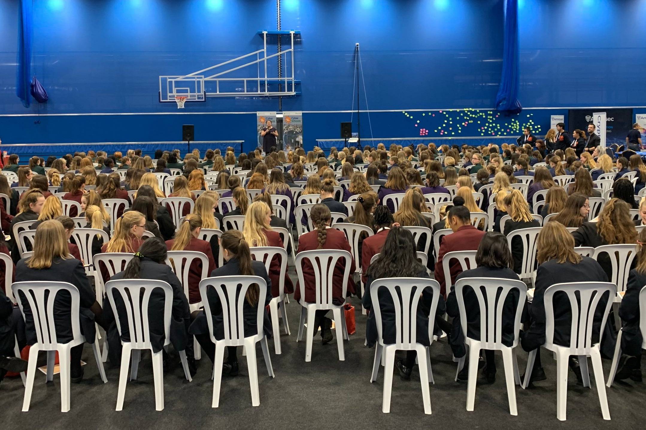 A big room with school girls sat in rows on white plastic chairs. The image is taken from the back of the room looking forward