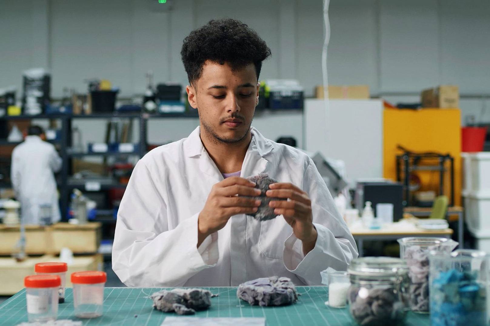 A mixed race man with black curly hair that is shaved at the sides. He is wearing a lab coat and holding a ball of dryer lint in warehouse setting