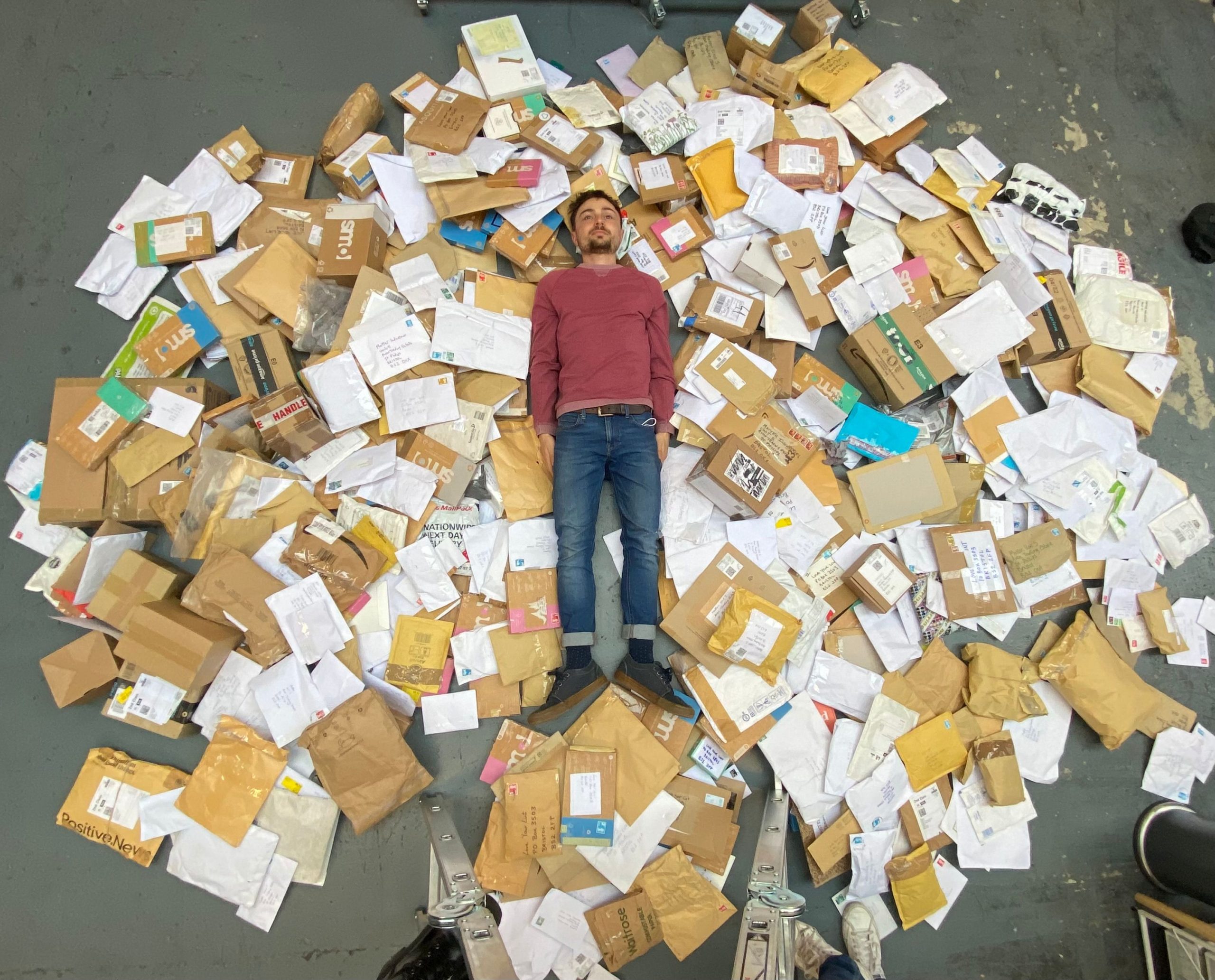 A white man with brown hair and facial hair, wearing a red jumper and blue jeans is lying down in a huge pile of different parcels. The shot is taken as a birds eye view