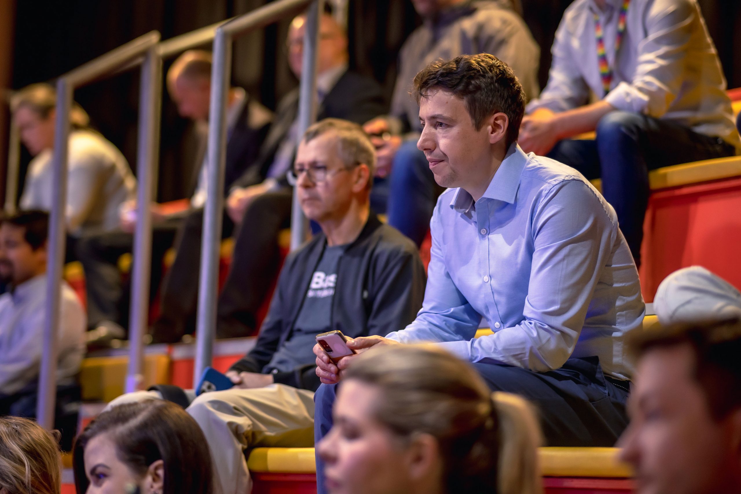 Central focus of the image is a white man with short brown hair sitting on the orange chairs, which have yellow cushions. He is wearing a light blue button up shirt and is leaning slightly forward with a phone in his hands rested on his knees. He is looking forward with a neutral expression. He is sat amongst several other people.
