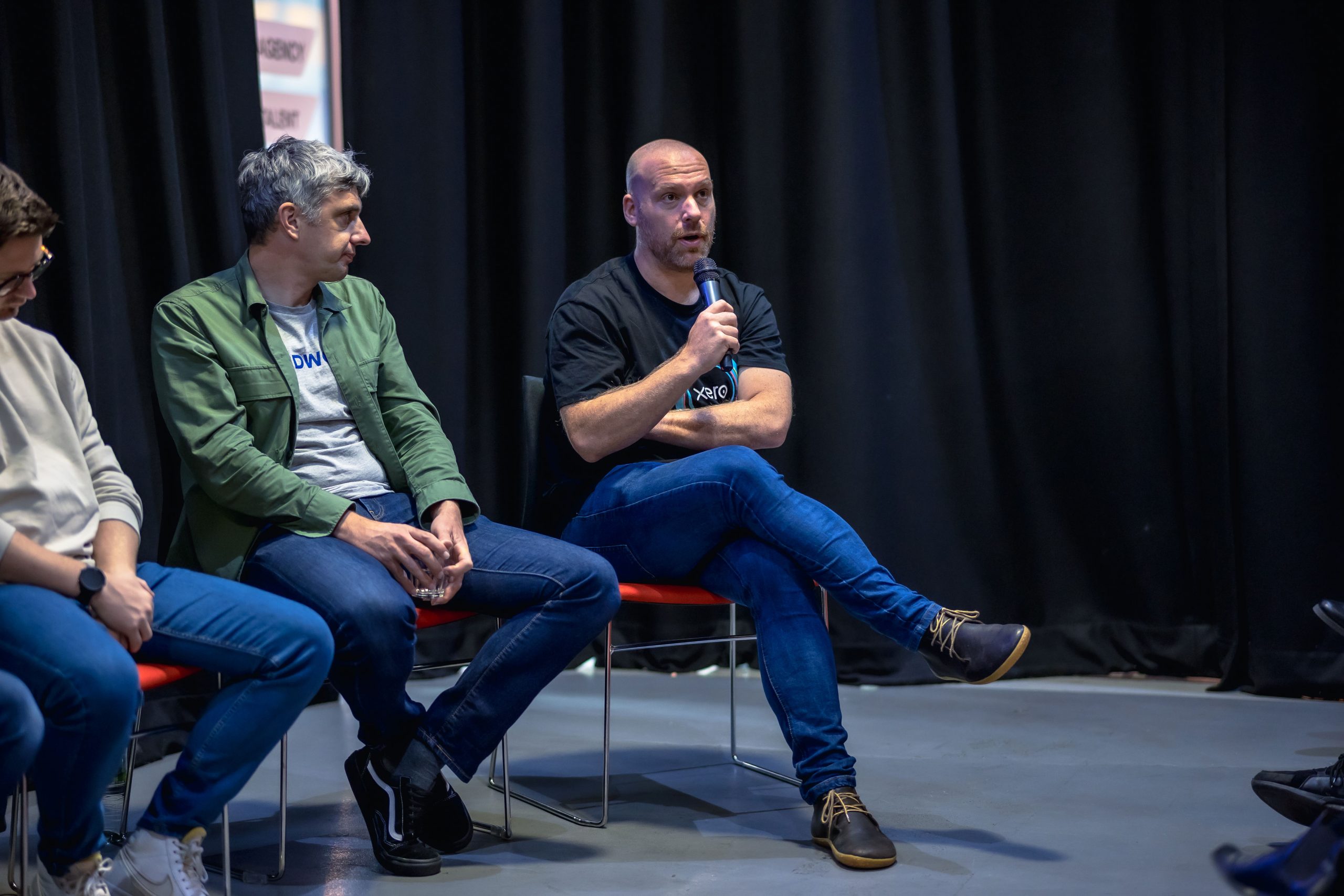 a bald white man with a light beard, wearing a black t-shirt and jeans. His legs are crossed and he is holding a mic, mid speaking. On his left is a white man with grey hair, jeans, a grey top and a khaki shirt/jacket. In the background is a floor length black curtain and grey flooring