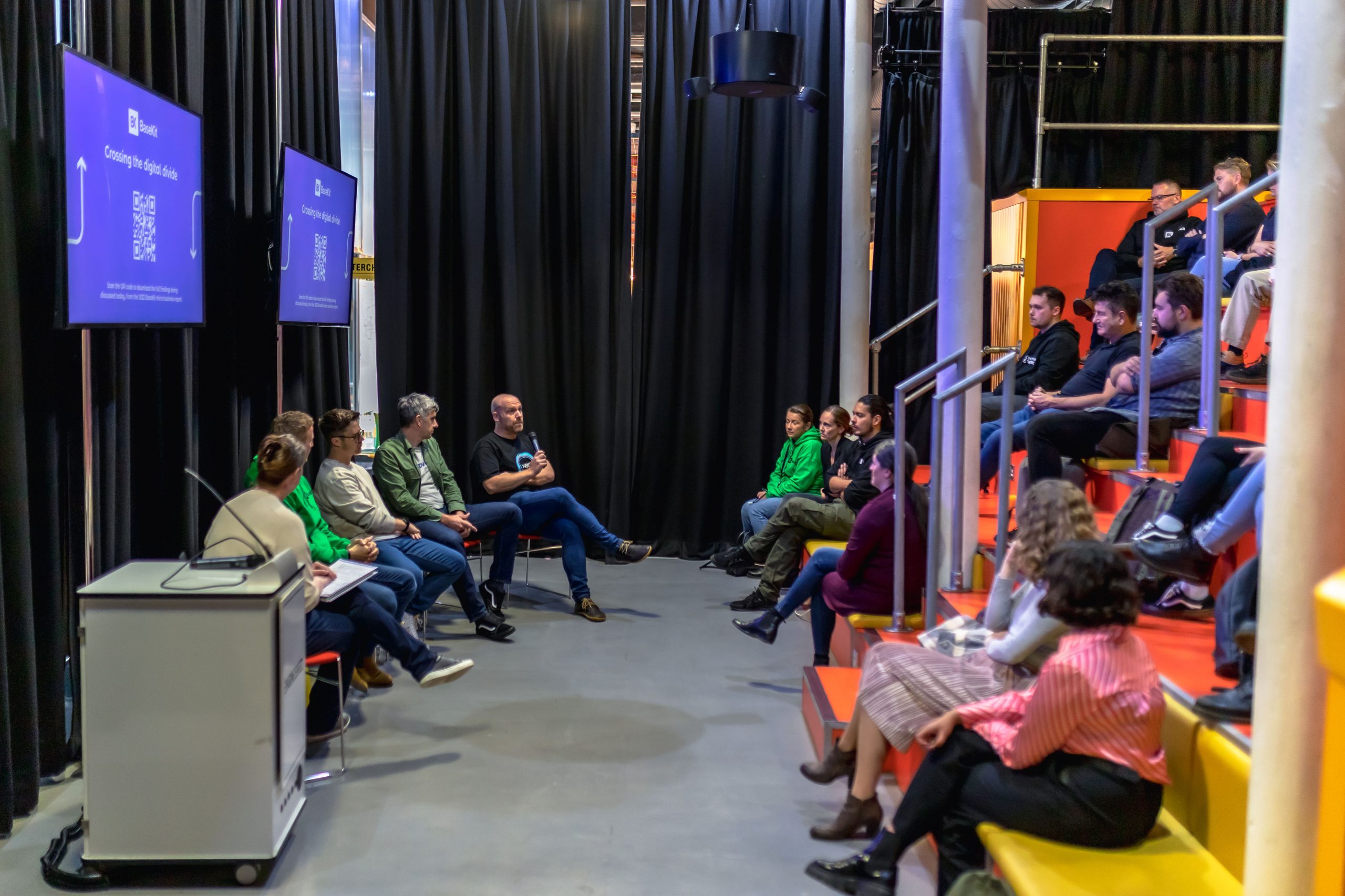 A side on view of the panel and audience. There is a grey floor, and around the sides is a long black floor length curtain. To the left, the panel of 5 is sitting in a row with two large screens behind and above them. On the screen is a purple background with white writing and a QR code and the event's title 