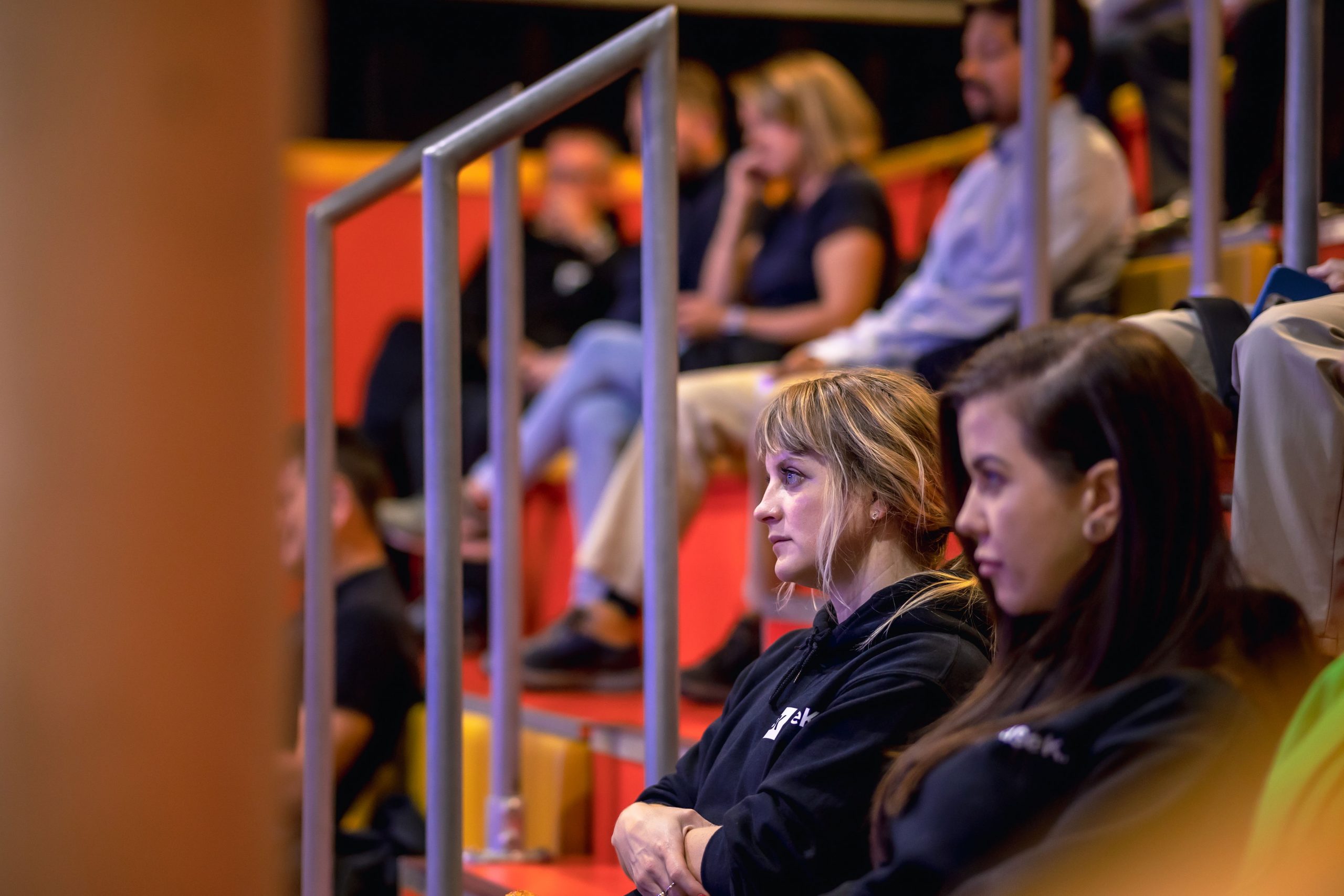 central focus is on a white woman with blonde hair. She has a fringe, the rest of her hair is tied back in a loose ponytail. She is wearing a black hoody and her arms are crossed, she is looking forward with a neutral expression. To her right is a white woman with straight dark hair, parted on the side and worn down. She is also wearing a black hoody. In the background is other people sat on the yellow and orange tiered benches. There are metal banisters in between the women and the rest of the audience.