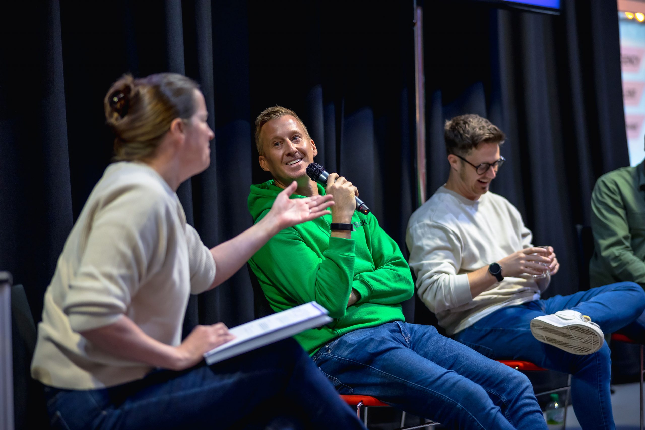 An image of a panel discussion - there are three people in the frame with a large black curtain behind them. From left to right, there is a white woman with brunette hair tied in a claw clip gesturing with her arm. She is wearing a beige long sleeve jumper with the sleeves pushed up to her elbows and blue denim jeans with a clipboard on her lap. To her right is a white man who has blonde short hair, wearing a bright green hoody and jeans, holding a mic and looking at the women whilst smiling. To his right is another white man with short brunette hair and black glasses, smiling looking down, wearing a beige crew neck jumper and jeans. He has his left leg crossed and resting his foot on his knee - he is wearing white trainers