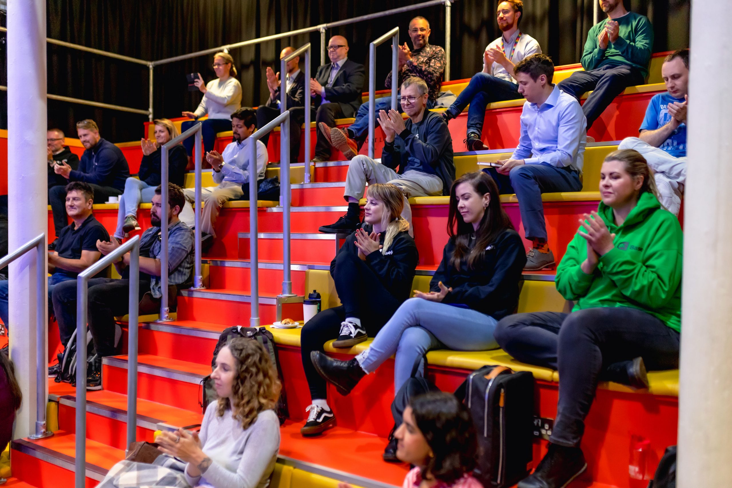 Audience of people sitting on a tiered bench across 7 levels, It has yellow cushioned seating and a hard orange back. Midway through there are stairs with a metal hand rail. At the top there is a black curtain