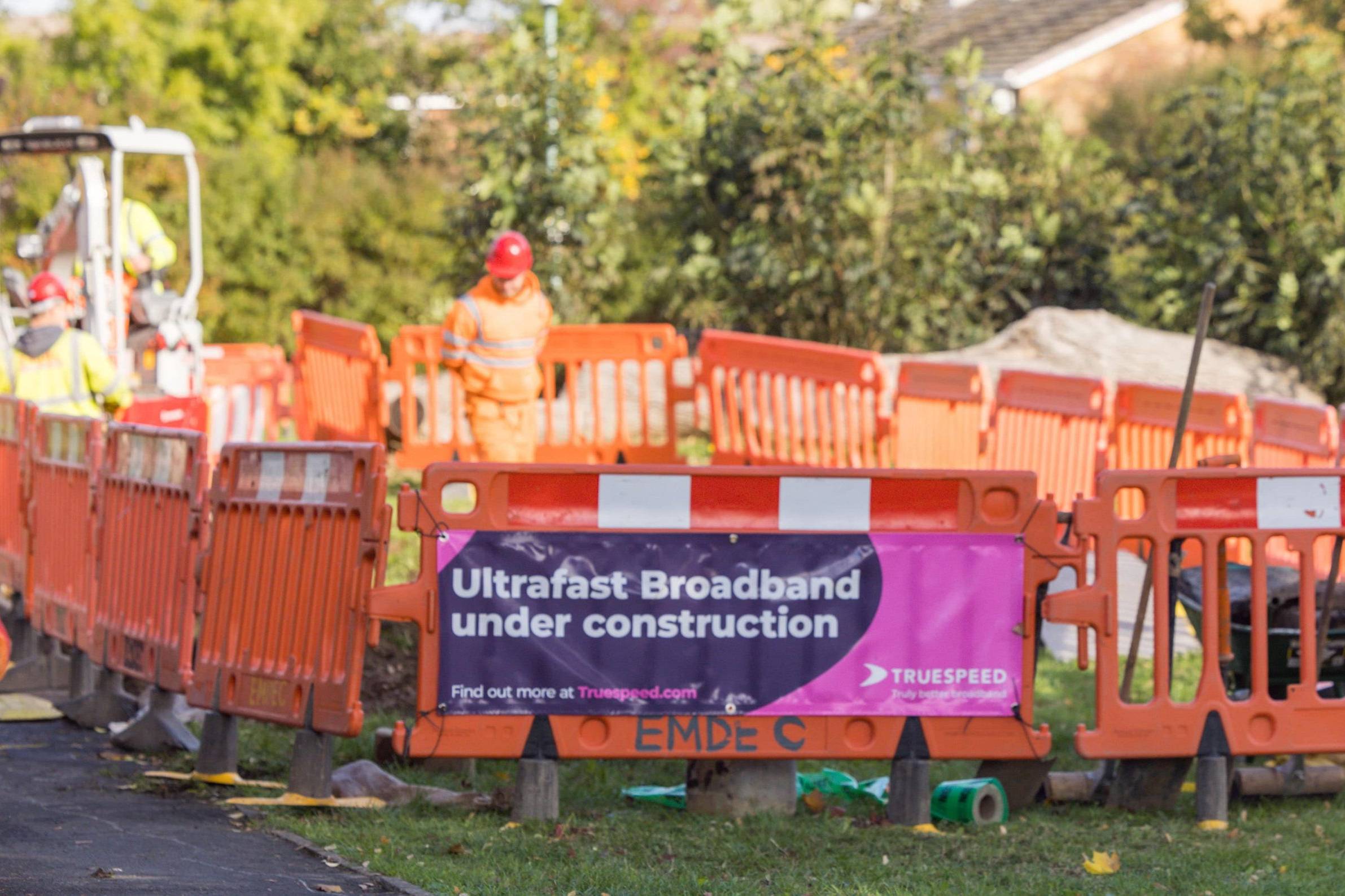At the foreground of the image there is orange plastic temporary fences with red and white tape along the top. These fences are positioned in a medium sized circular shape. It is a construction site with a construction worker wearing orange high vis and a red helmet stood in the middle, blurred in the background. On his left, there is a 360 excavator operator digging machine, controlled by someone wearing yellow high vis. On the nearest fence, there is a pink and purple banner which says