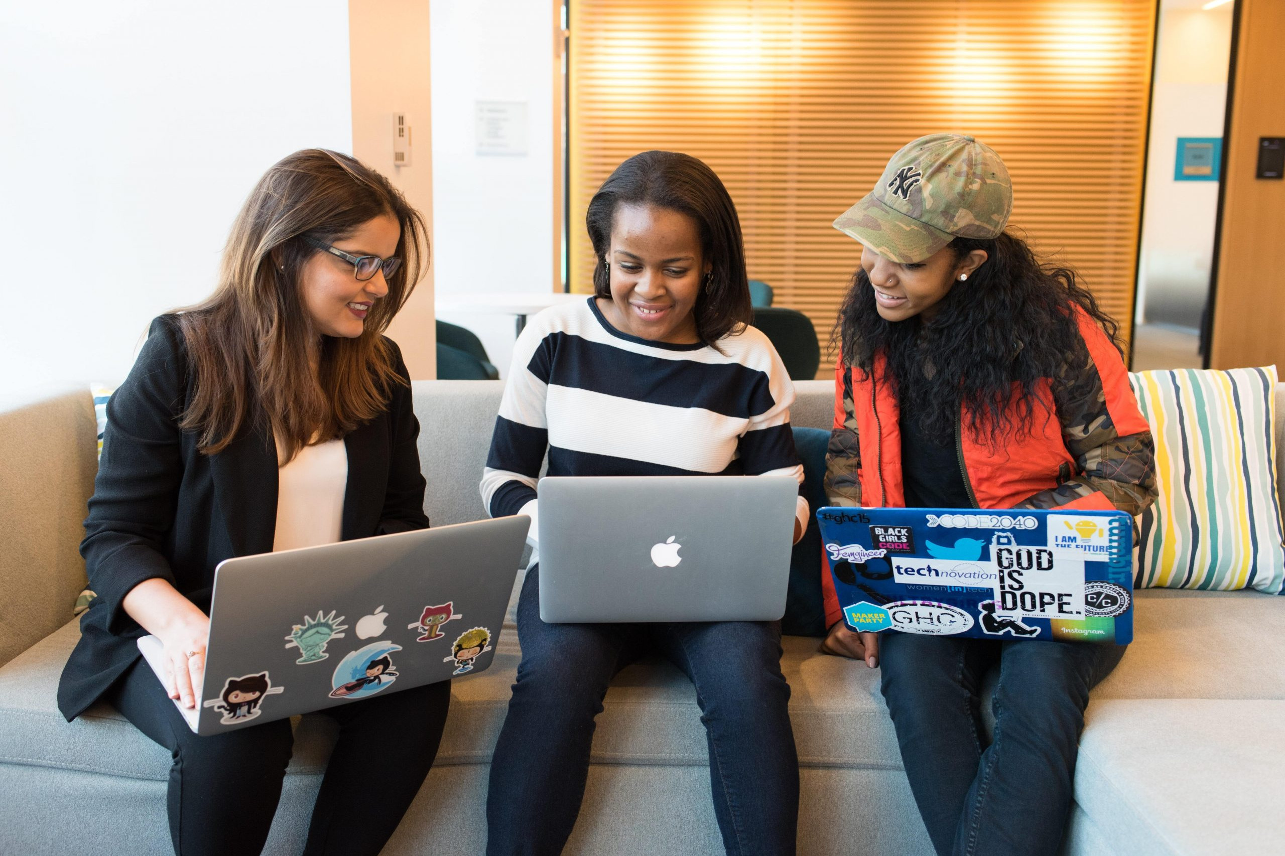 Three Black women sat on a sofa each with a laptop on their laps, smiling at one another
