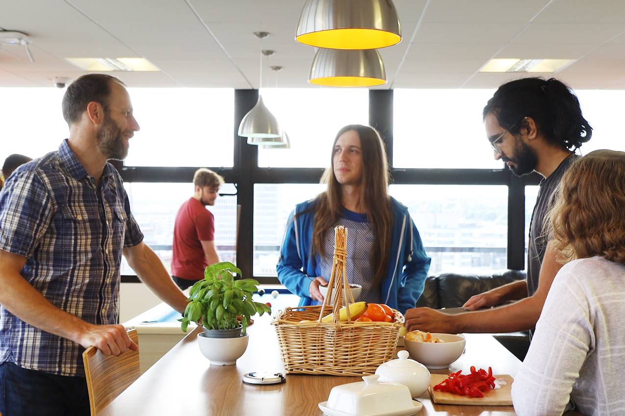 A group of people are congregated around a breakfast bar at work, conversing and preparing food