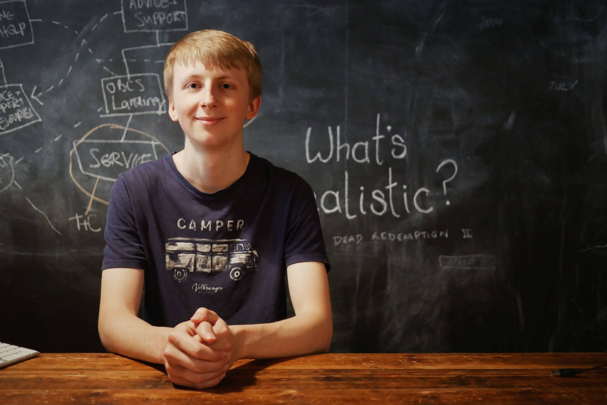 Torchbox Academy participant. Young man sits at a wooden desk with a black board behind him
